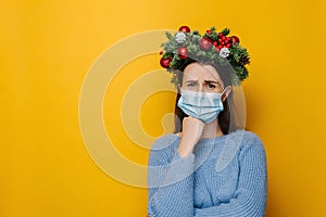 Unhappy young female wearing medical face mask, looks sad, shrugs with crossed arms, wears spruce holiday wreath around head