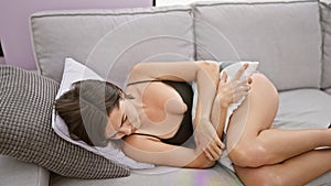 Unhappy young hispanic woman with short hair, suffering stomach ache, worriedly touching her belly, lying on sofa in living room