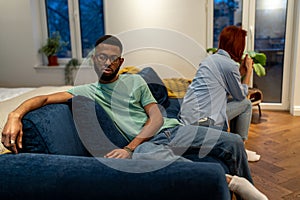 Unhappy young interracial couple sit separately on sofa at home not talking after fight