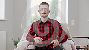 Unhappy young man in eyeglasses losing in video game leaning back on armchair. Portrait of sad Caucasian male gamer