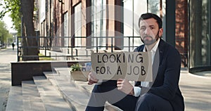 Unhappy young man showing looking for job carton banner near office building. Dismissed male worker in suit searching