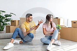 Unhappy young multiracial couple having argument, sitting on floor in their new apartment on moving day