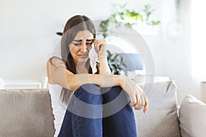 Unhappy young woman covering face with hands, crying alone close up, depressed girl sitting on couch at home, health problem or