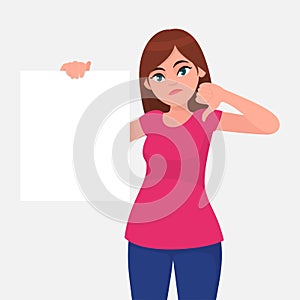 Unhappy young woman holding a blank / empty sheet of white paper or board and gesturing thumbs down sign.