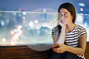 Unhappy young woman holding a smartphone in the evening cityscape