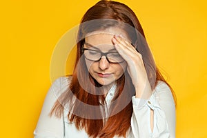 Unhappy young woman with red long hair and glasses suffers a headache, migraine on a yellow wall background. Stress