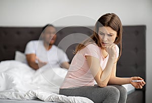 Unhappy young woman sitting on bed with pregnancy test, black husband screaming at her, indoors