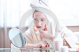 Unhappy young woman with a towel on her head detects acne on her face. Concept of hygiene and care for the skin