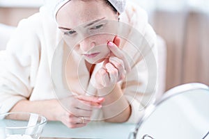 Unhappy young woman with a towel on her head detects acne on her face. Concept of hygiene and care for the skin