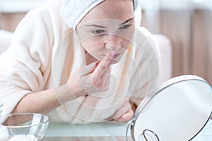 Unhappy young woman with a towel on her head detects acne on her face. Concept of hygiene and care for the skin