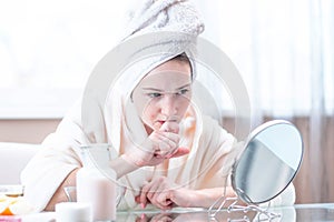 Unhappy young woman with a towel on her head detects acne on her face. Concept of hygiene and care for the skin