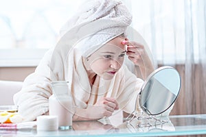 Unhappy young woman with a towel on her head detects acne on her face. Concept of hygiene and care for the skin