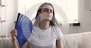 Unhappy young woman waving fan feeling uncomfortable hot at home