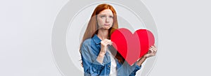 Unhapy brokenhearted cute redhead girl, sulking and looking up uneasy, distressed holding big red valentines day heart