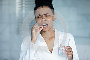 Unhealthy african woman holding glass of water takes painkiller