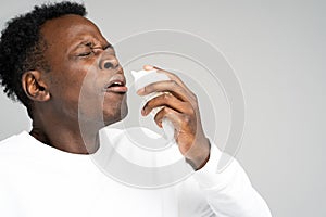 Unhealthy Afro-American man blowing nose and sneeze into tissue or napkin, experiences allergy symptoms, closed eyes, standing