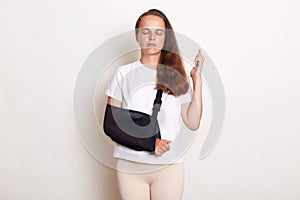 Unhealthy arm broken Caucasian woman with arm sling supported on her hand, being injured, keeps eyes closed and crossed fingers,