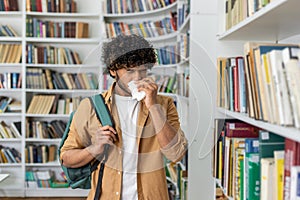 Unhealthy man in casualwear using paper napkin while sneezing and standing by aisle with collection of novels. Sick