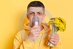 Unhealthy man wearing casual style hoodie posing isolated over yellow background, holding bouquet of dandelions and breathing with