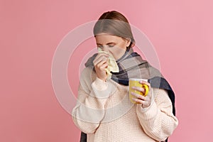 Unhealthy sick ill blond woman wrapped in scarf standing with cup of tea, suffering from runny nose.