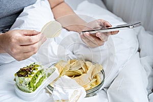 unhealthy woman eating junk food salty snack potato chips and dessert , cake in bed