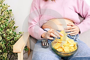 Unhealthy woman eating junk  potato chips in bowl hand holding big, fat belly sitting on sofa watching tv