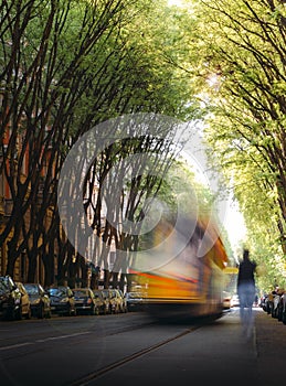Unidentifable person running towards an out of blur tram on a tree-lined path on the street