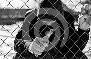 Unidentifiable teenage boy behind wired fence holding a paper knife at correctional institute, focus on the fence in black and whi