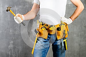 unidentified handyman standing with a tool belt with construction tools and holding a hammer against grey background. DIY tools