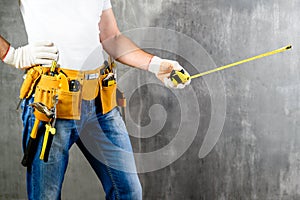 unidentified handyman standing with a tool belt with construction tools and holding roulette against grey background. DIY tools