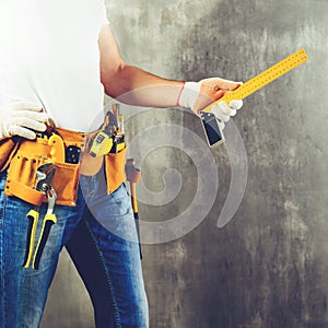 unidentified handyman standing with a tool belt with construction tools and holding roulette against grey background, toned image