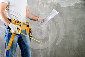 unidentified handyman standing with a tool belt with construction tools and holding roulette and project plan against grey