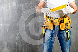 unidentified handyman standing with a tool belt with construction tools and holding a ruler against grey background. DIY tools