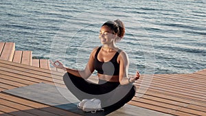 Unity with nature. Young african american woman athlete meditating near water, sitting in lotus position on pier