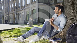 University student sitting under tree on campus, solving task in notebook, smart