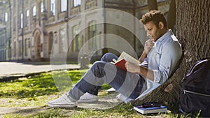 University student sitting under tree reading book with gripping plot, engrossed