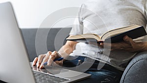 University student using laptop or computer for studying or learning book, reading textbook at home. Knowledge and skill training