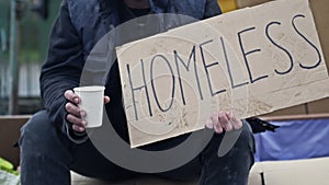 An unkempt-looking man sits with a begging cup and holds a handwritten HOMELESS poster. Hearty passers-by throw coins