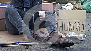 An unkempt-looking man sits with a begging cup and holds a handwritten HUNGRY poster. Hearty passers-by throw coins into