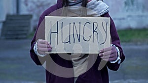 An unkempt-looking young woman sits with a begging cup and holds a handwritten HUNGRY poster.