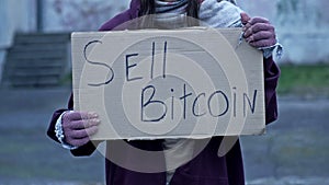 An unkempt-looking young woman sits with a begging cup and holds a handwritten SELL BITCOIN poster.