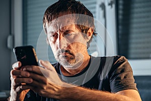 Unkempt man using cell phone indoors, alone adult caucasian male holding smartphone