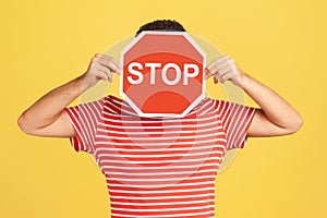 Unknown anonymous man in red striped t-shirt hiding his face behind red stop sign board, scared victim of humiliation and violence