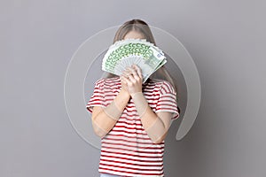 Unknown anonymous rich little girl wearing striped T-shirt hiding her face behind euro banknotes.