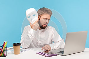 Unknown bearded man putting of white face mask sitting at workplace an looking at camera with mysterious facial expression, hiding