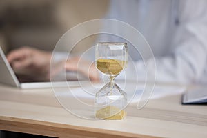 Unknown businesswoman working on laptop, focus on hourglass on desk