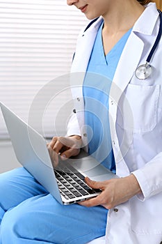 Unknown female doctor typing on laptop computer while sitting at chair in clinic. Medical staff, compulsory work concep