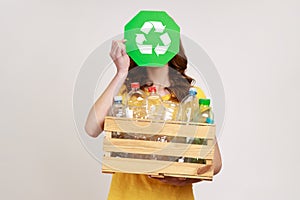 Unknown female holding wooden box full of plastic bottles and covering face with recycle sing