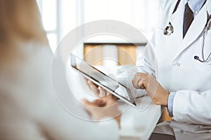 Unknown male doctor and patient woman discussing current health examination while sitting in clinic and using tablet