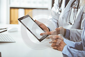 Unknown male doctor and patient woman discussing current health examination while sitting in clinic and using tablet